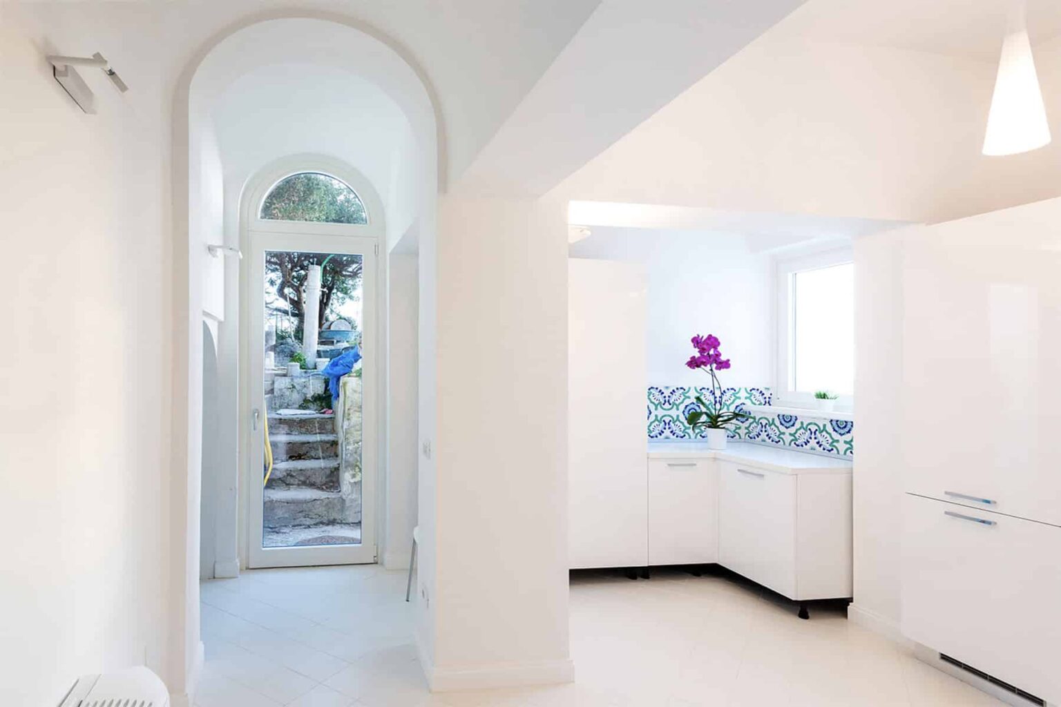 Bright and modern kitchen with white cabinetry and colorful tile backsplash, featuring an arched entrance leading to a garden view. | Only on SopranoVillas
