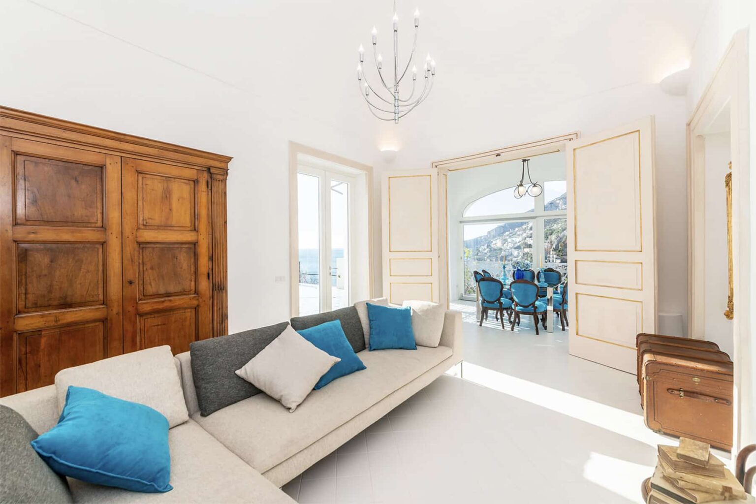 Bright and modern living room featuring a stylish gray sofa with blue accent pillows, wooden cabinetry, and large doors leading to a dining area with a scenic view. Ideal for showcasing contemporary interior design. | Only on SopranoVillas