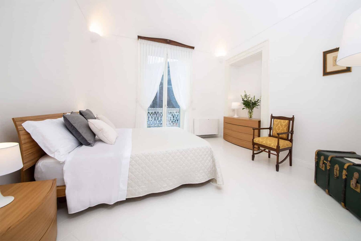 Bright and airy modern bedroom featuring a comfortable bed, elegant wooden furniture, and natural light streaming through a window. The decor includes a cozy chair and a stylish dresser, creating a serene and inviting atmosphere. Ideal for relaxation and comfort. | Only on SopranoVillas