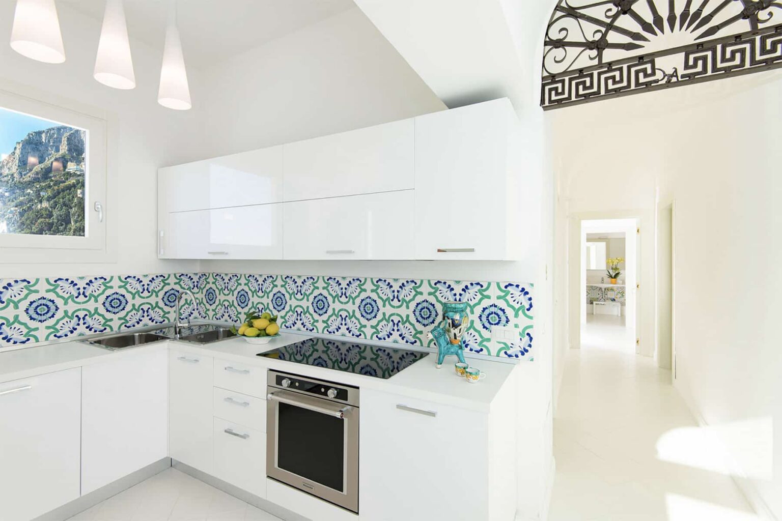 Modern kitchen design featuring white cabinetry, a stylish blue and green patterned backsplash, stainless steel appliances, and natural light streaming through a window. | Only on SopranoVillas