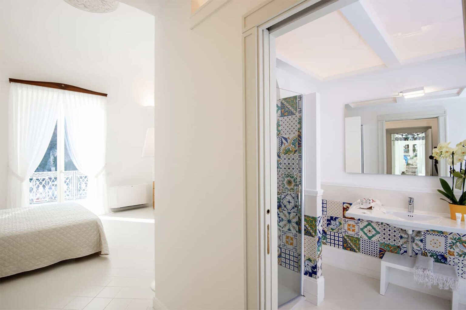 Bright and airy bedroom leading to a stylish bathroom with colorful tiled accents and modern fixtures, showcasing a serene and elegant interior design. | Only on SopranoVillas
