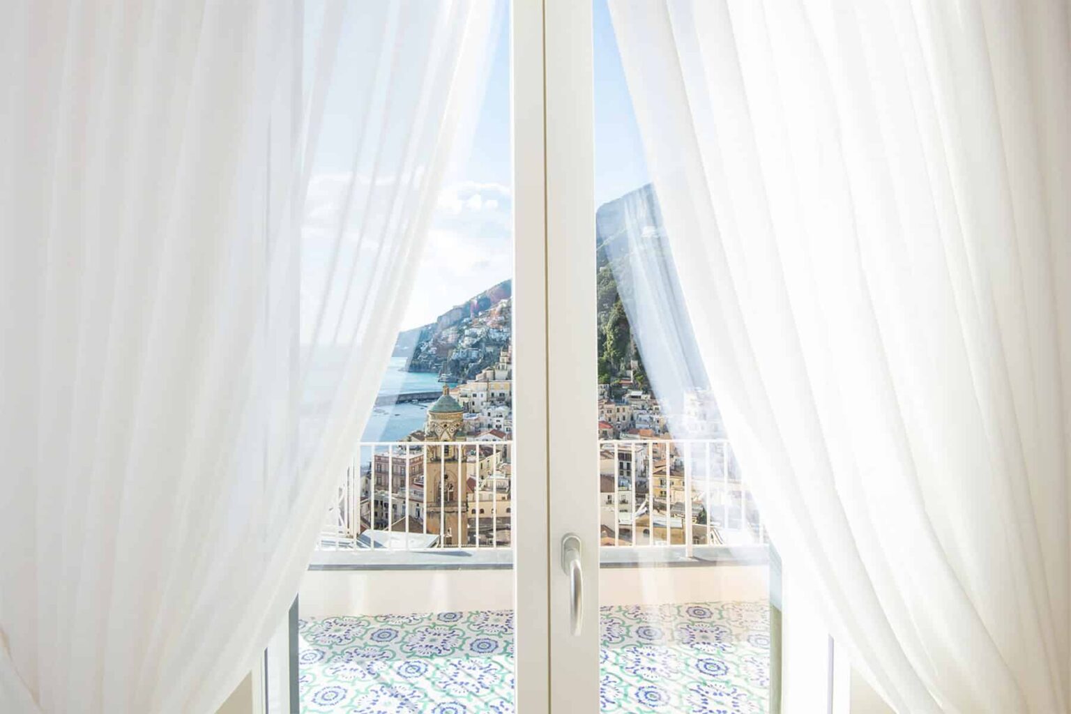 Bright and airy view through white sheer curtains, revealing a scenic landscape with mountains and a body of water beyond a balcony. Ideal for showcasing a serene and inviting atmosphere in home decor or travel content. | Only on SopranoVillas