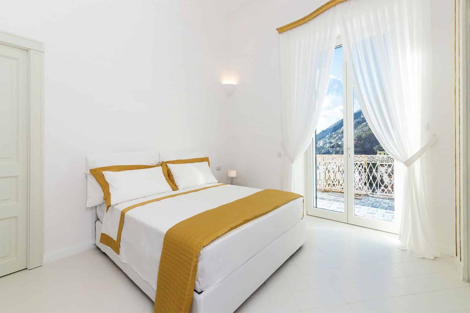 Cozy bedroom featuring a white bed with orange accents, large windows with sheer curtains, and a modern, minimalist design. Natural light illuminates the space, enhancing its inviting atmosphere. | Only on SopranoVillas