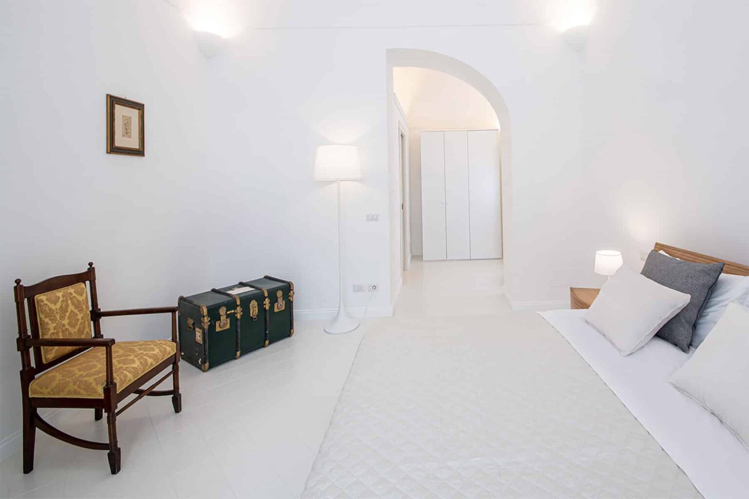 Bright and airy bedroom featuring a white color palette, a comfortable bed with decorative pillows, an elegant wooden chair, and a vintage trunk. The room includes soft lighting and a minimalist design, creating a serene atmosphere. | Only on SopranoVillas