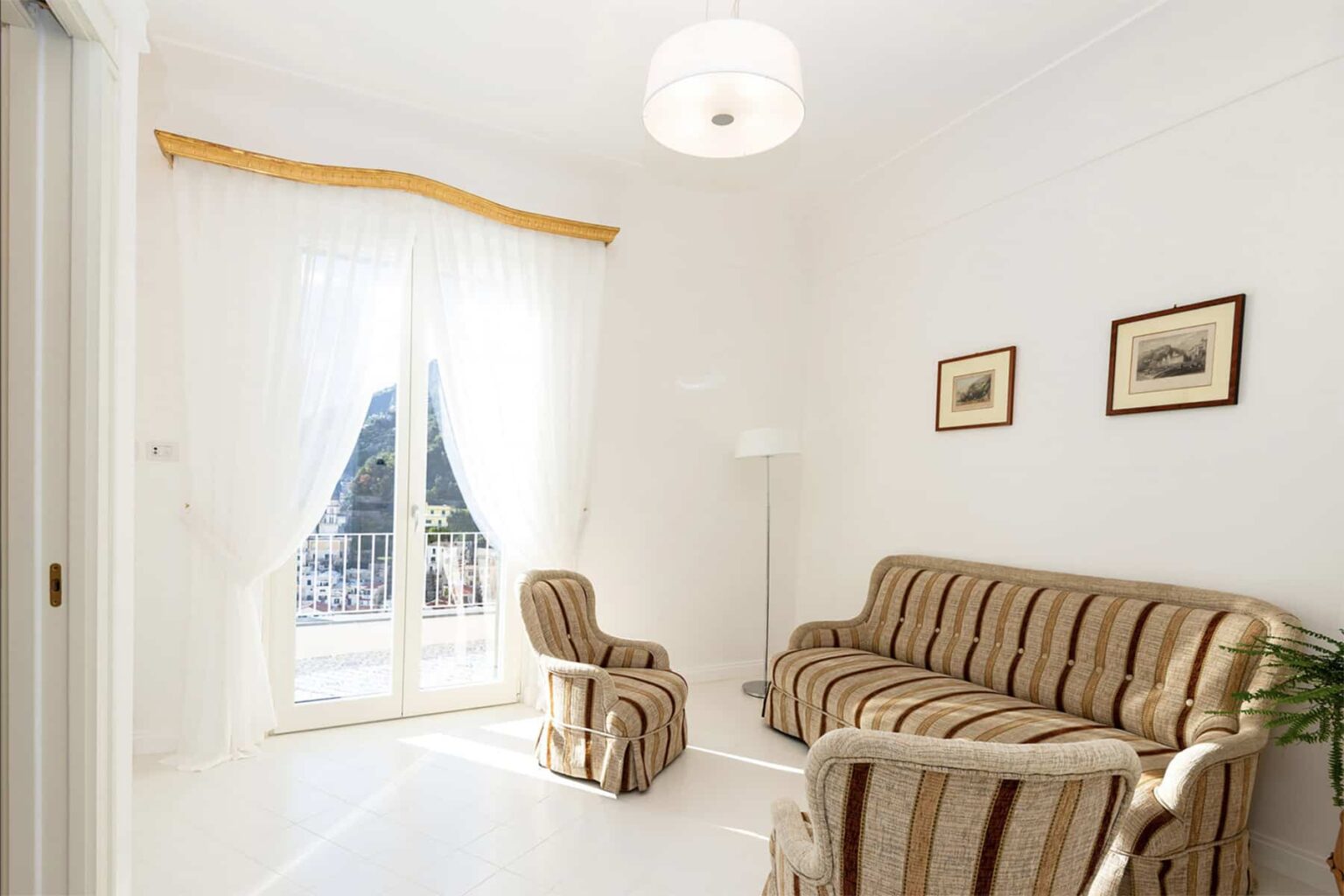 Bright and airy living room featuring a striped sofa, armchair, and large windows with sheer curtains, enhancing the natural light and spacious feel. Decorated with framed artwork, the room exudes a cozy and inviting atmosphere. | Only on SopranoVillas