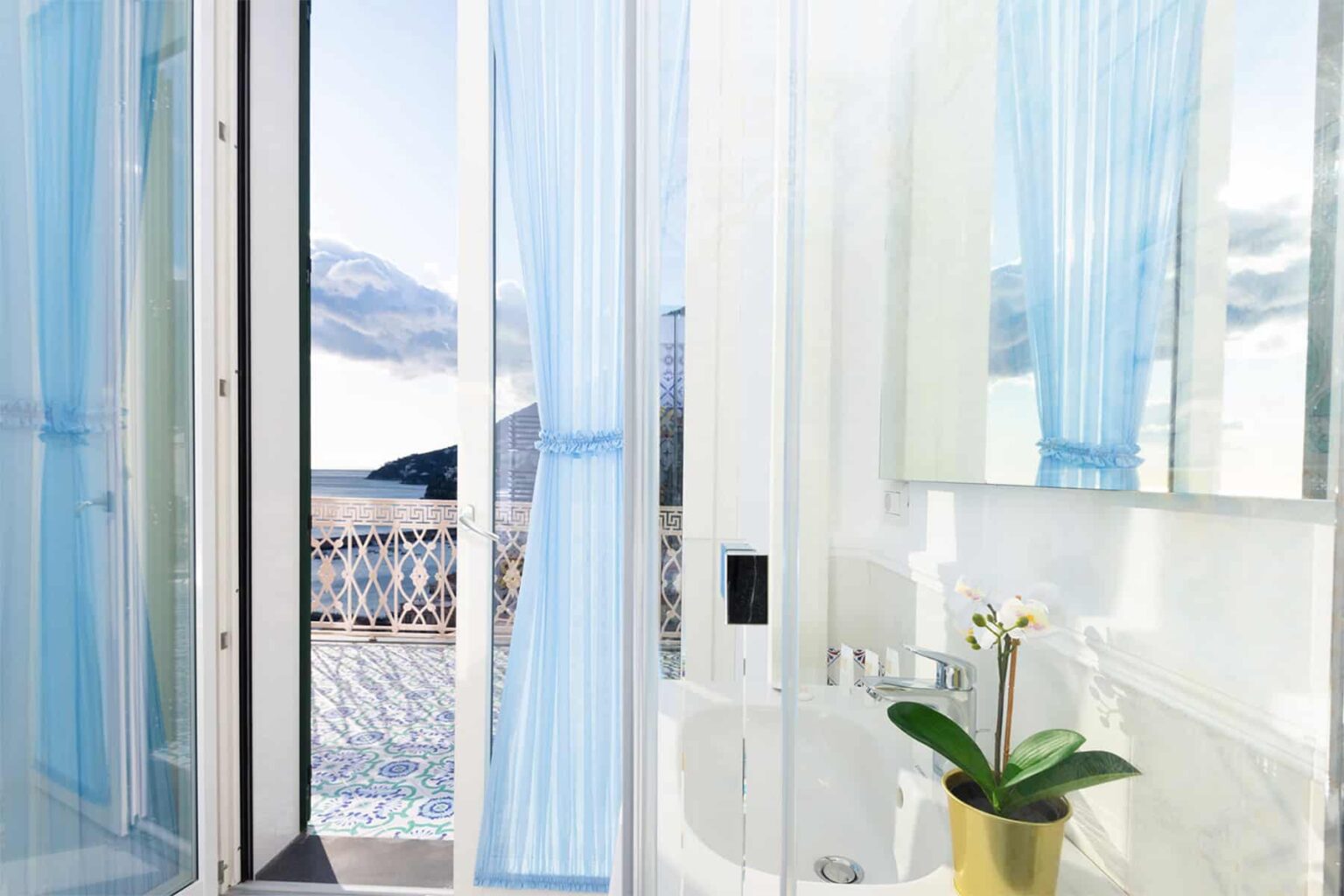 Bright and airy bathroom featuring large windows with sheer blue curtains, a modern mirror, and a decorative potted orchid, overlooking a scenic outdoor view. | Only on SopranoVillas