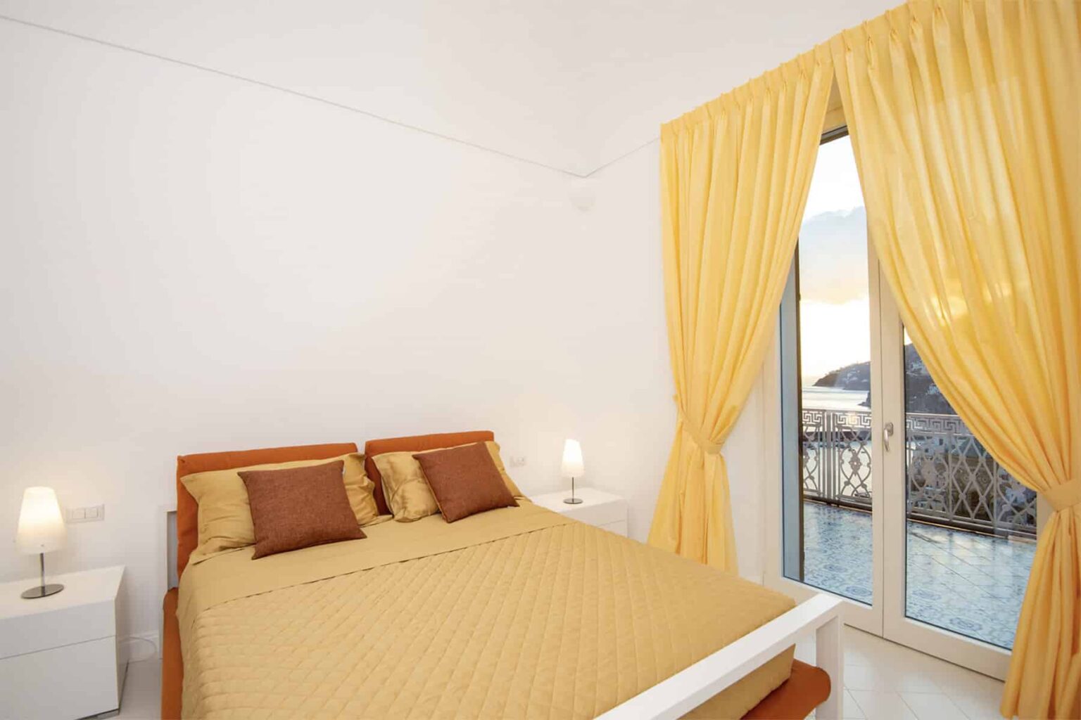 Cozy bedroom featuring a queen-sized bed with yellow bedding and decorative pillows, complemented by bright yellow curtains that frame a large window with a scenic view. The room is well-lit with bedside lamps and a minimalist design, creating a warm and inviting atmosphere. | Only on SopranoVillas