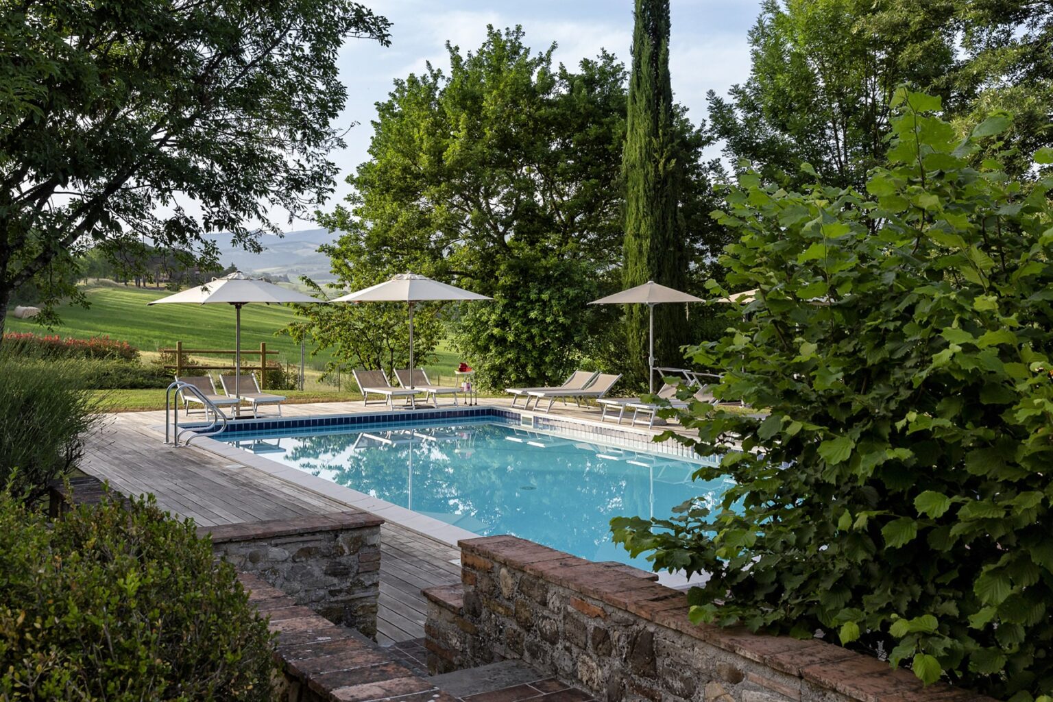 Serene outdoor swimming pool surrounded by lush greenery and trees, featuring sun loungers and umbrellas, with a picturesque landscape in the background. | Only on SopranoVillas