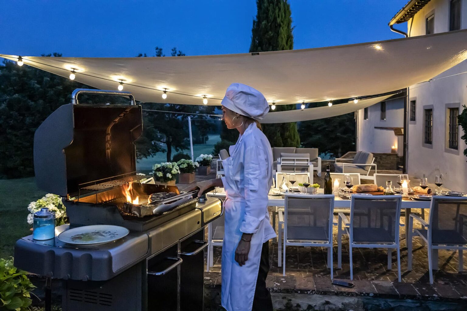Chef grilling outdoors at night under a canopy with string lights, surrounded by a beautifully set dining area. | Only on SopranoVillas
