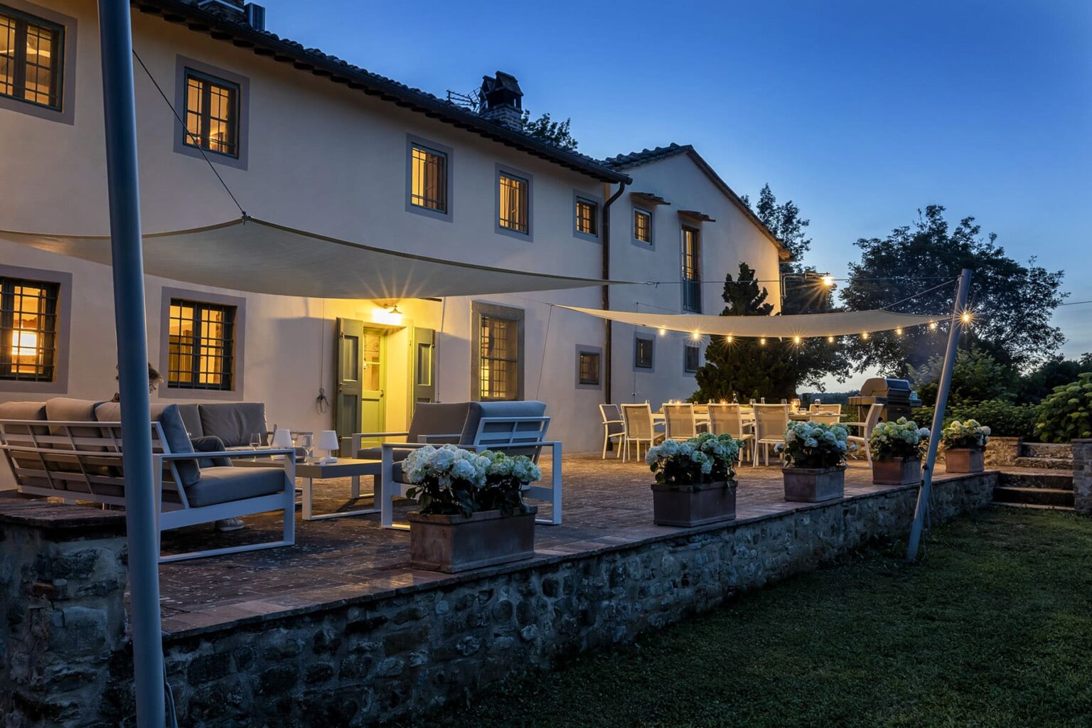 Charming countryside villa at dusk with outdoor seating, illuminated windows, and a spacious patio adorned with potted flowers, perfect for relaxation and gatherings. | Only on SopranoVillas