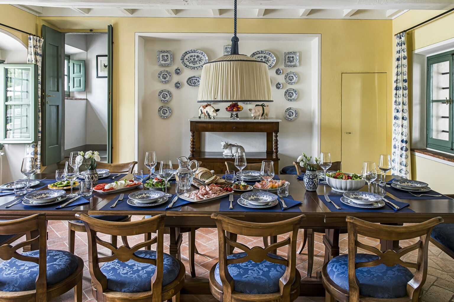 Elegant dining room featuring a large wooden table set for a meal, adorned with blue tableware and fresh food. The walls are painted yellow and decorated with decorative plates, while large windows allow natural light to fill the space. A stylish chandelier hangs above the table, creating a warm and inviting atmosphere. | Only on SopranoVillas