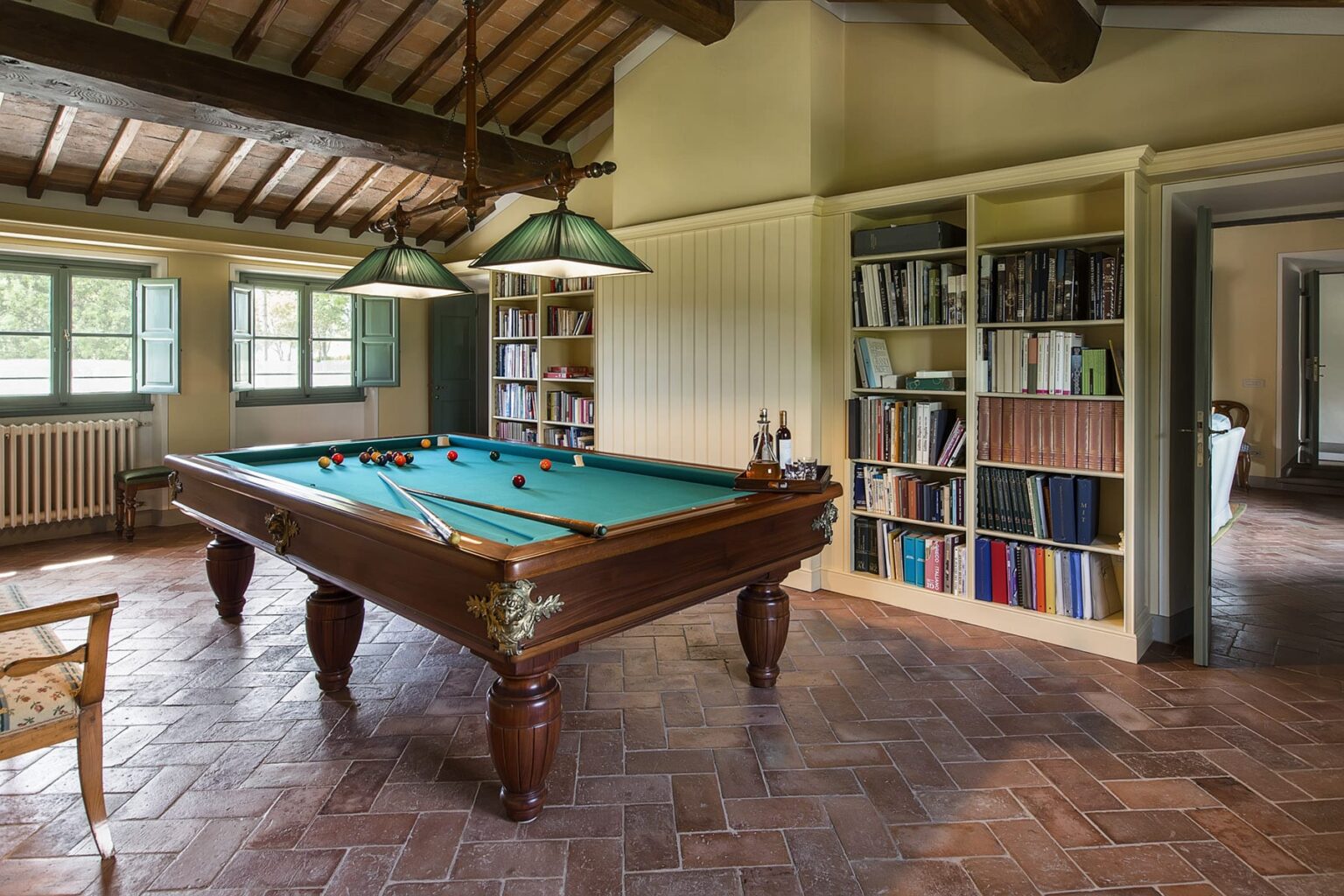 Interior view of a game room featuring a classic pool table with green felt, surrounded by a cozy atmosphere, wooden beams, and a bookshelf filled with books. Natural light enters through large windows, enhancing the inviting space. | Only on SopranoVillas