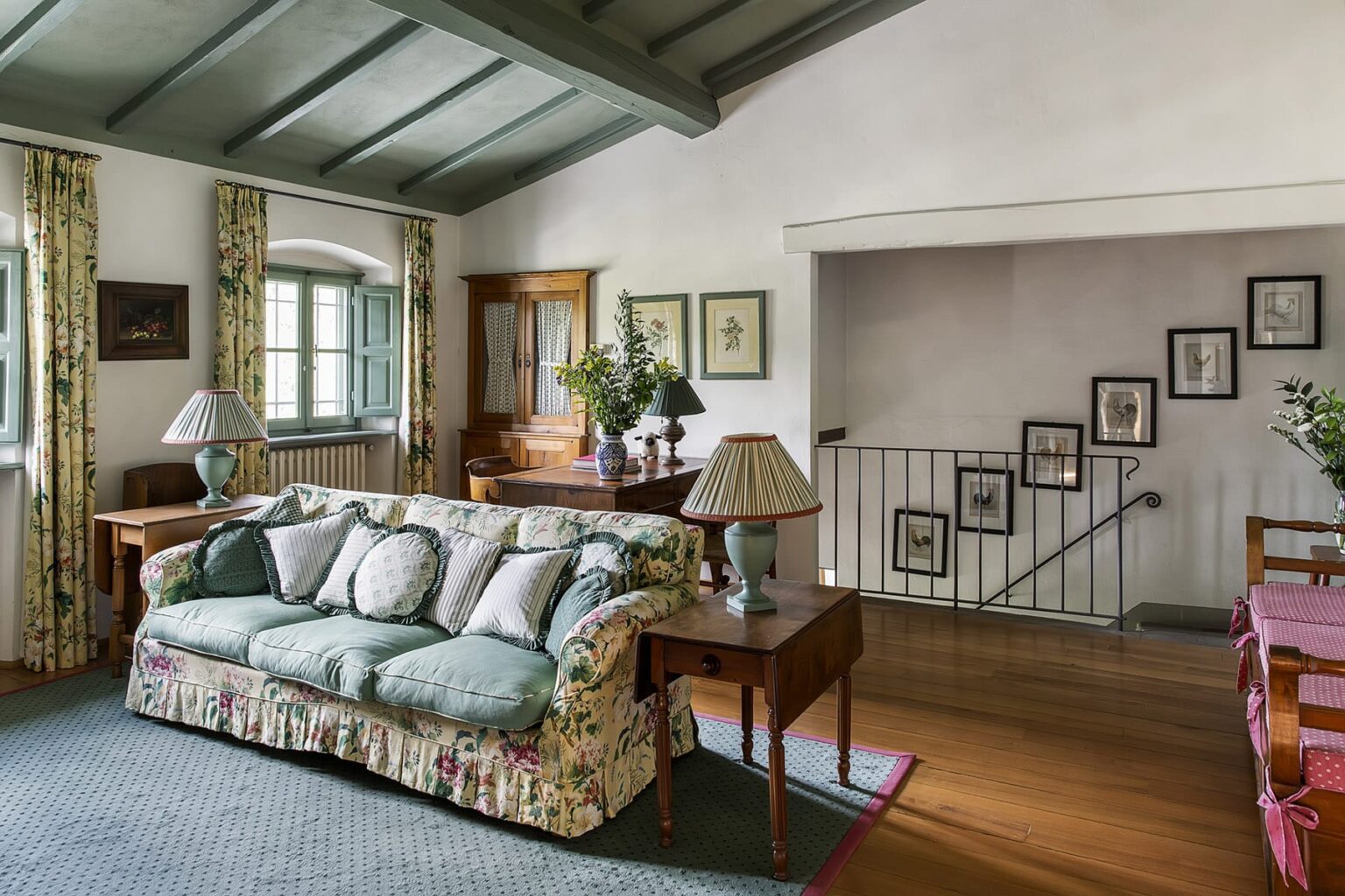 Cozy living room featuring a floral-patterned sofa, wooden furniture, and decorative lamps, with large windows and a staircase in the background. The room has a warm ambiance enhanced by natural light and tasteful decor. | Only on SopranoVillas