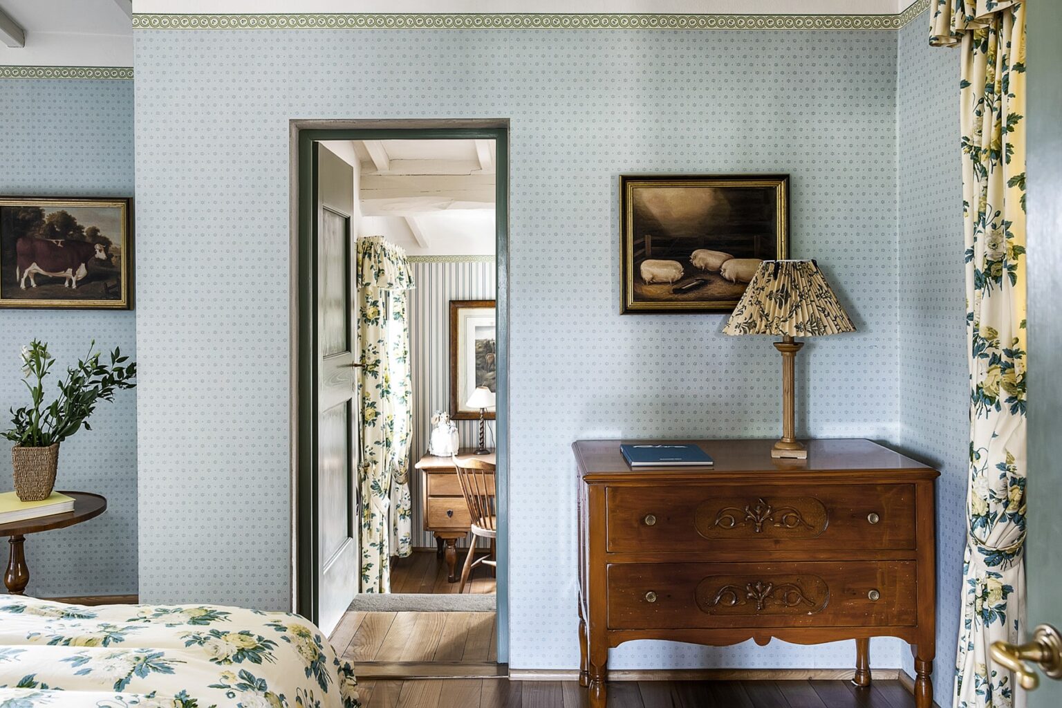 Cozy interior of a vintage-style bedroom featuring soft blue walls, a wooden dresser, a decorative lamp, and floral curtains. A doorway leads to an adjoining room, enhancing the inviting atmosphere. | Only on SopranoVillas