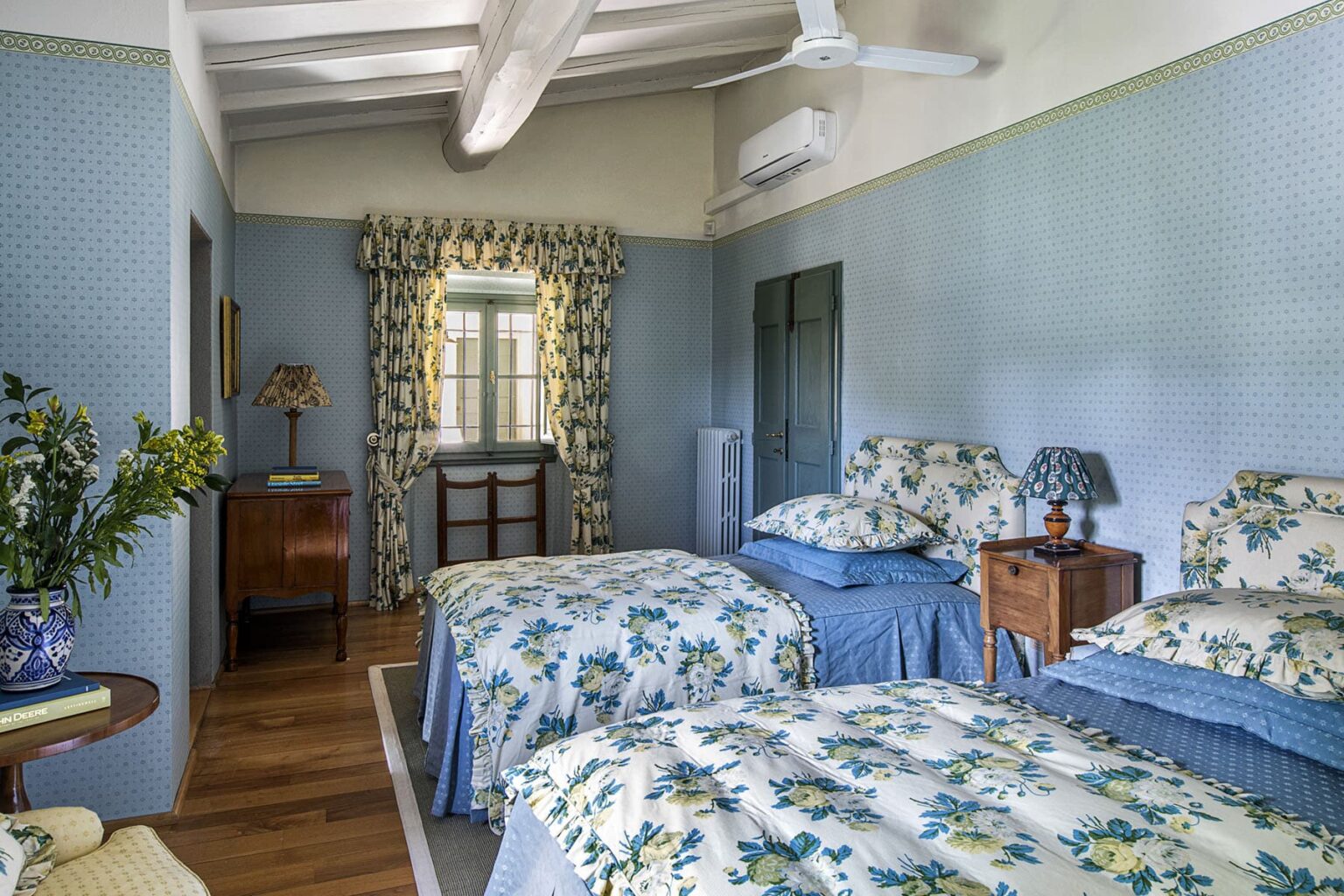 Cozy guest room featuring two twin beds with floral bedding, blue walls, and natural light from a window. Decor includes a vintage bedside table, a lamp, and floral curtains, creating a charming and inviting atmosphere. | Only on SopranoVillas