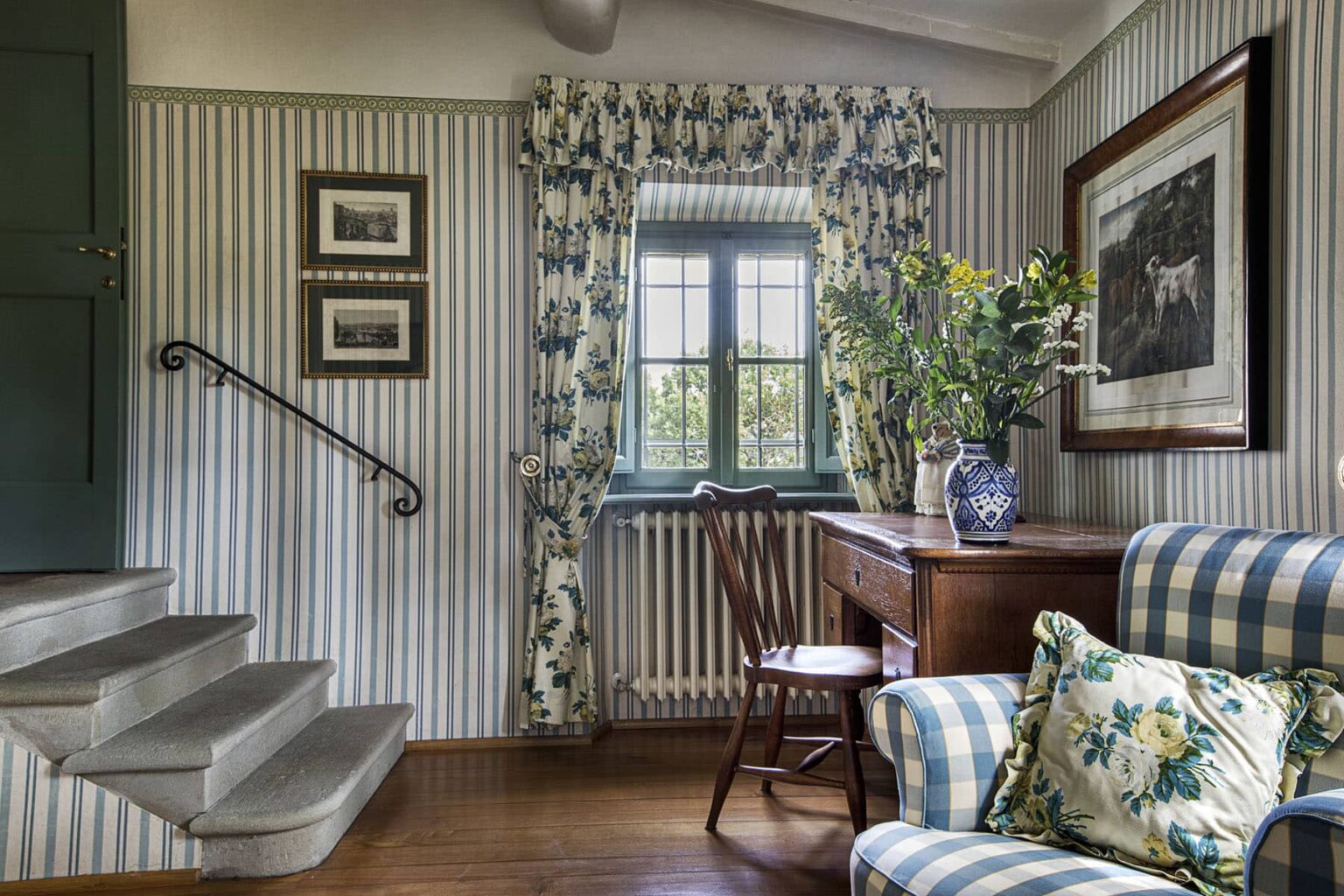 Cozy interior of a stylish living room featuring striped wallpaper, a wooden staircase, a vintage chair, a desk with floral decor, and a window adorned with patterned curtains. | Only on SopranoVillas
