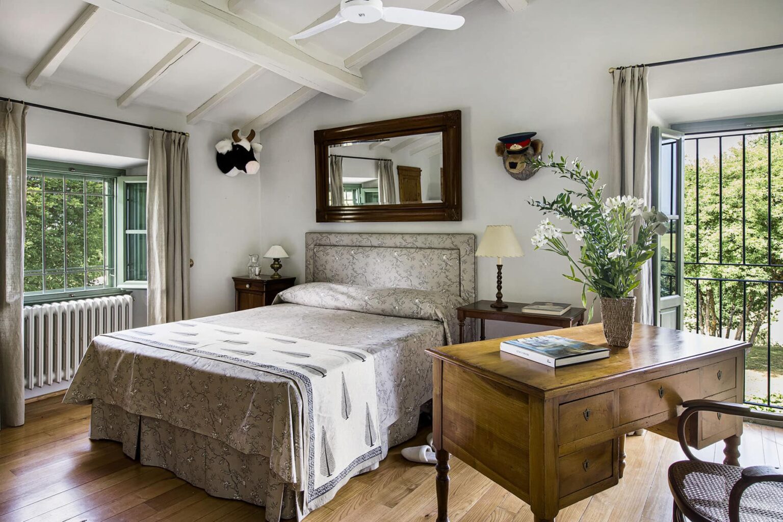 Cozy bedroom featuring a double bed with a decorative quilt, wooden desk with a chair, and large windows allowing natural light. The room is adorned with a mirror and wall decor, creating a warm and inviting atmosphere. | Only on SopranoVillas