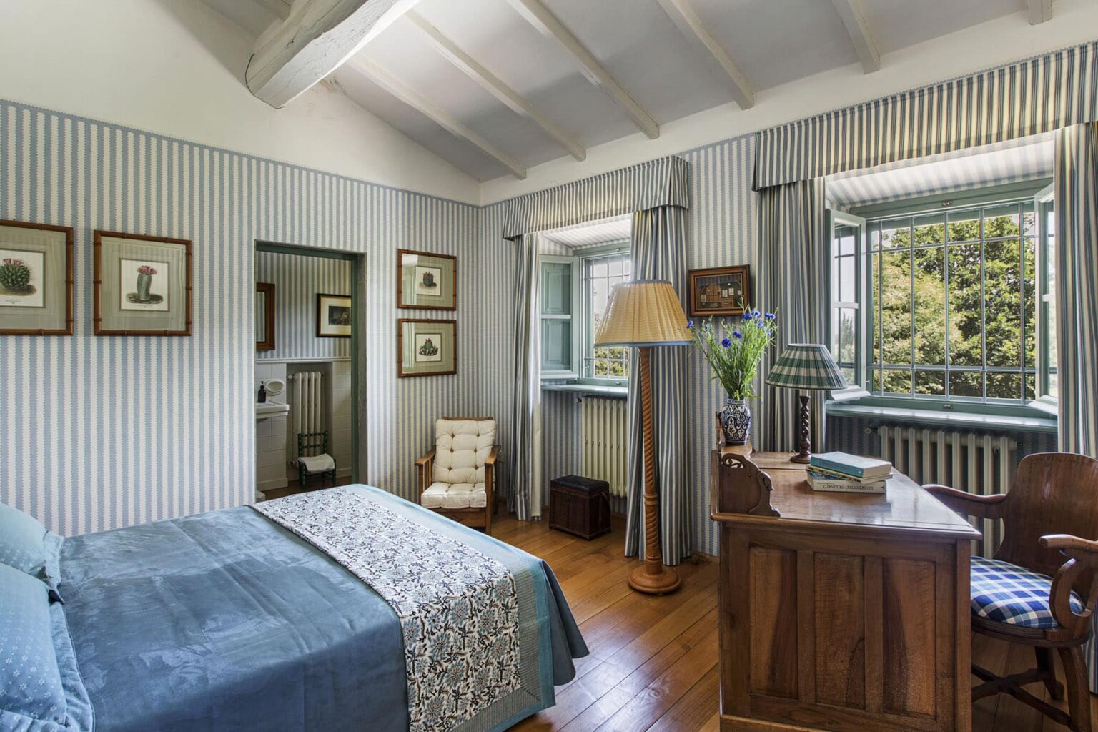 Cozy bedroom interior featuring a blue bedspread, wooden desk, and vintage decor, with large windows allowing natural light and scenic views. | Only on SopranoVillas