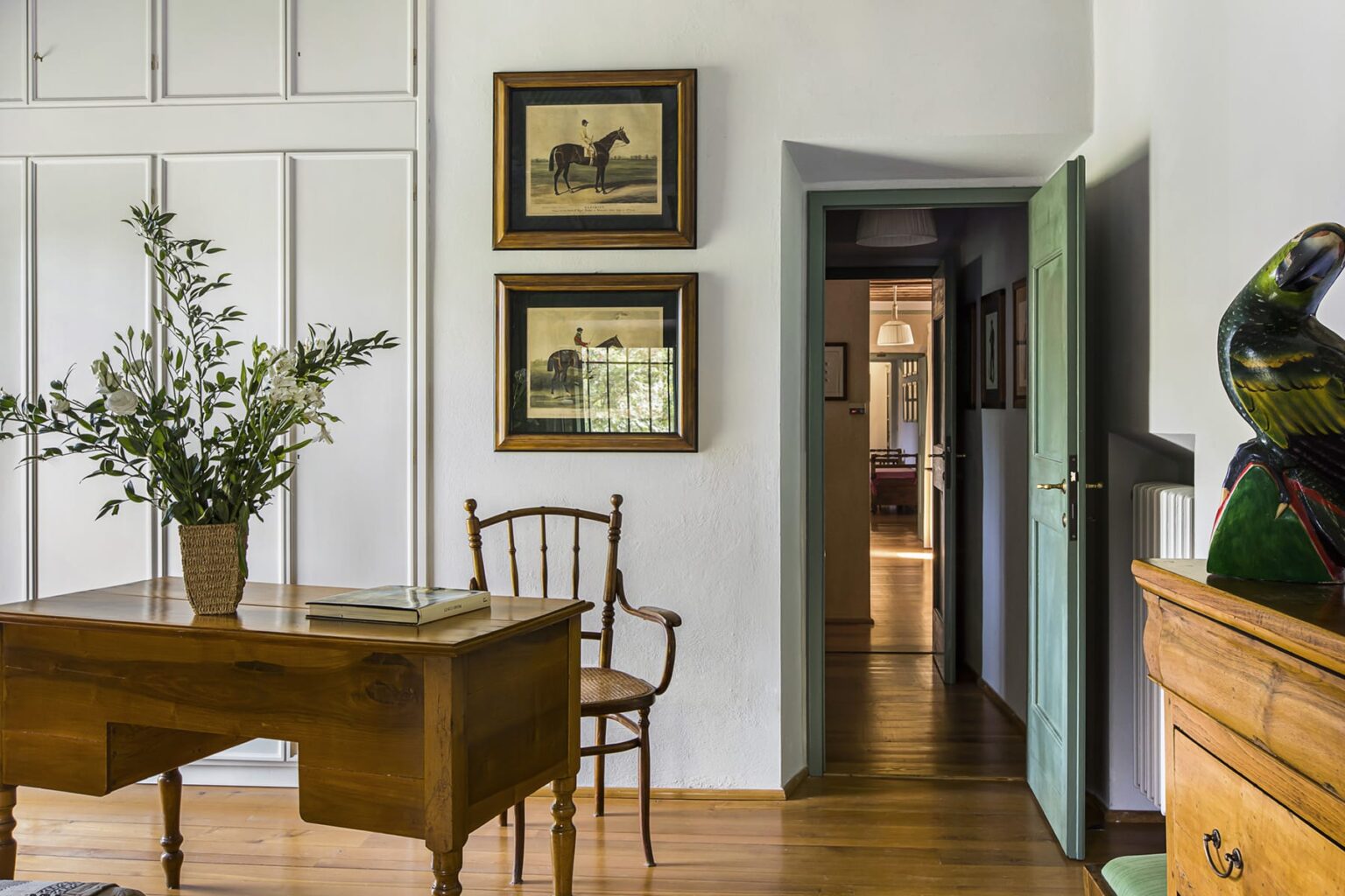 Cozy interior of a home office featuring a wooden desk, a chair, and framed artwork on the walls. A doorway leads to another room, with natural light illuminating the space. Greenery in a vase adds a touch of nature to the decor. | Only on SopranoVillas