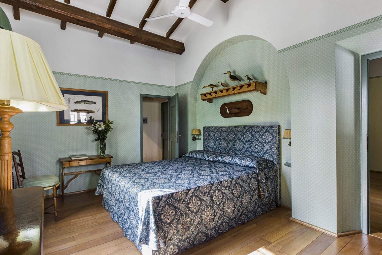 Cozy bedroom featuring a blue patterned bedspread, wooden ceiling beams, and a small desk with a vase of flowers, ideal for relaxation and comfort. | Only on SopranoVillas