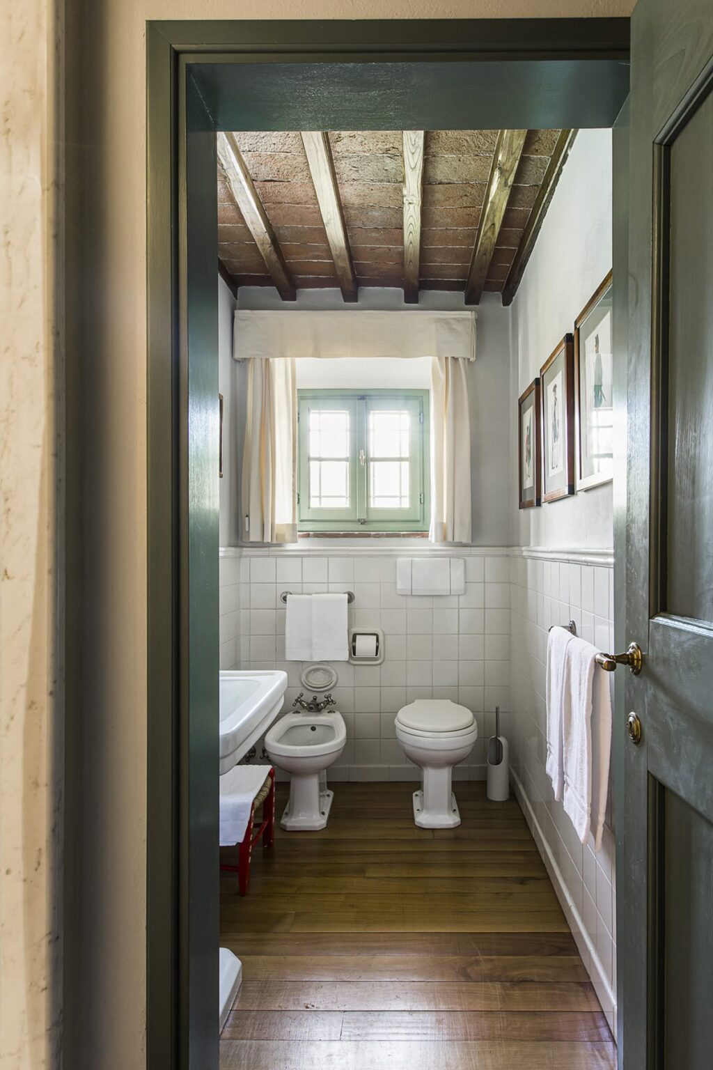 Small, modern bathroom featuring a white toilet, sink, and tiled walls. Natural light enters through a green-framed window, complemented by white curtains. Two towels hang on the wall, adding to the clean and inviting atmosphere. | Only on SopranoVillas