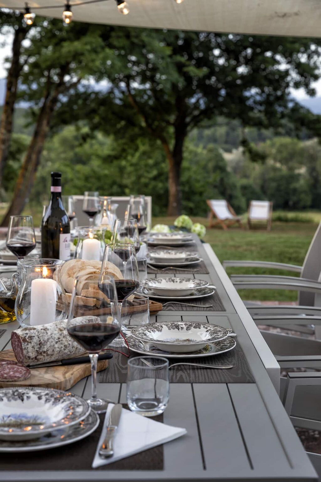 Elegant outdoor dining setup featuring a long table with wine glasses, a bottle of red wine, candles, and freshly baked bread, surrounded by lush greenery and lounge chairs in the background. | Only on SopranoVillas