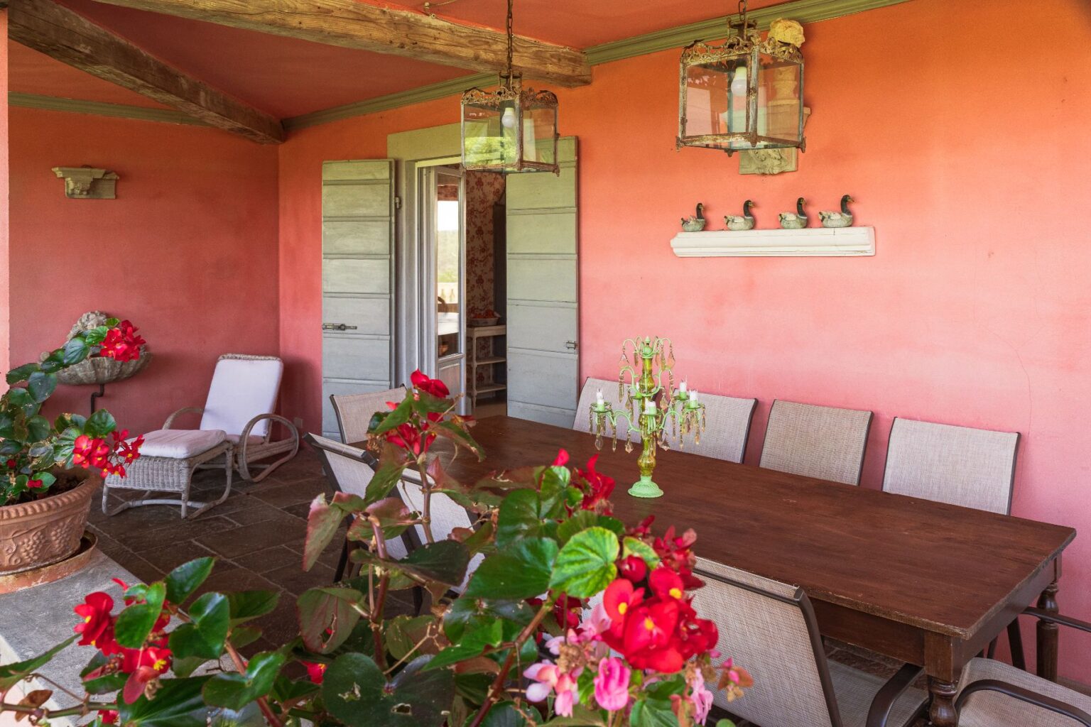 Cozy outdoor dining area with a wooden table and chairs, surrounded by vibrant flowers and a warm coral wall. Features include hanging lanterns and decorative accents, creating an inviting atmosphere for gatherings. | Only on SopranoVillas