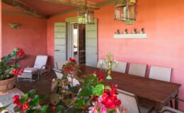 Cozy outdoor dining area with a wooden table and chairs, surrounded by vibrant flowers and a warm coral wall. Features include hanging lanterns and decorative accents, creating an inviting atmosphere for gatherings.