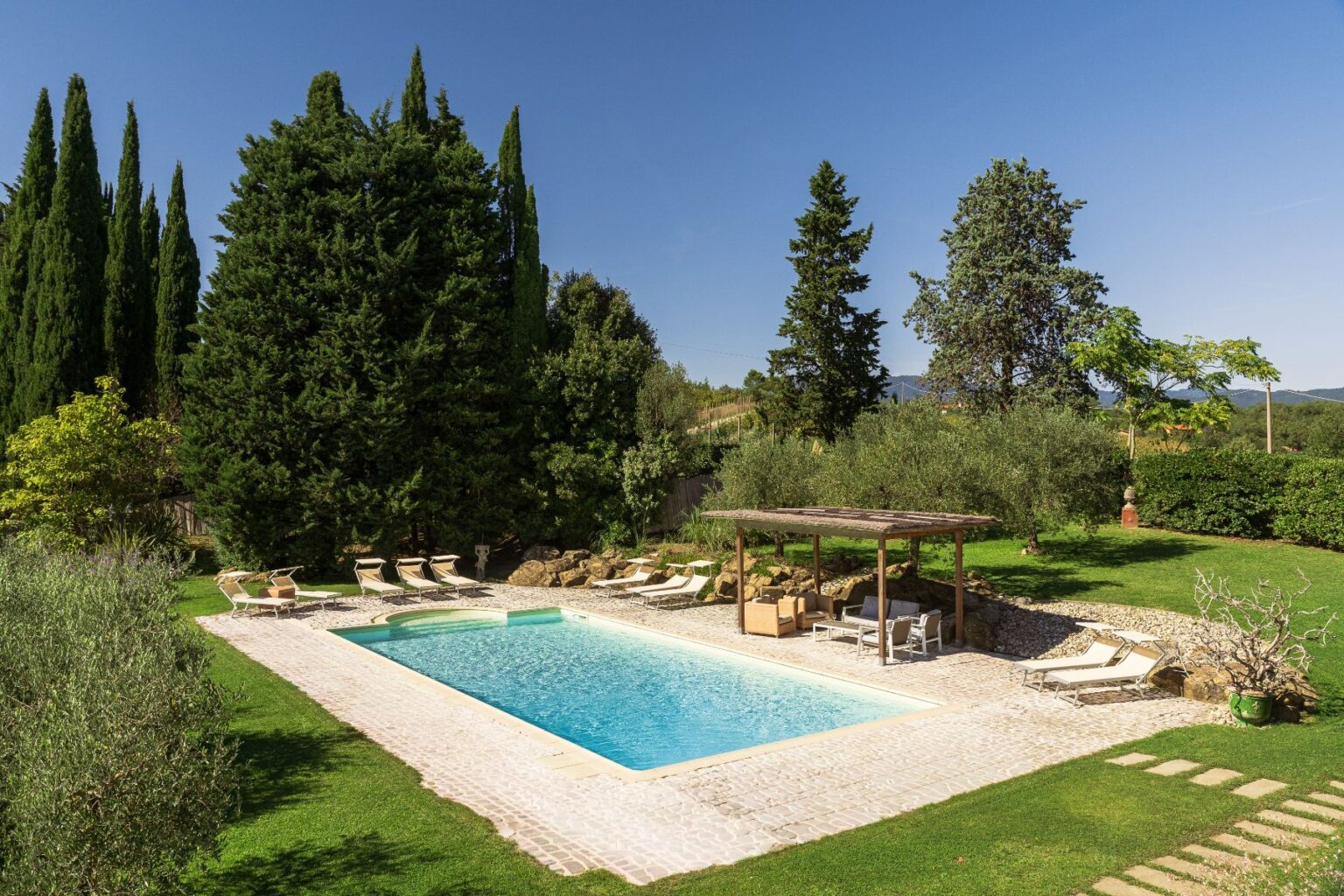 Luxurious outdoor pool surrounded by lush greenery, sun loungers, and a shaded seating area, ideal for relaxation and leisure in a serene garden setting. | Only on SopranoVillas