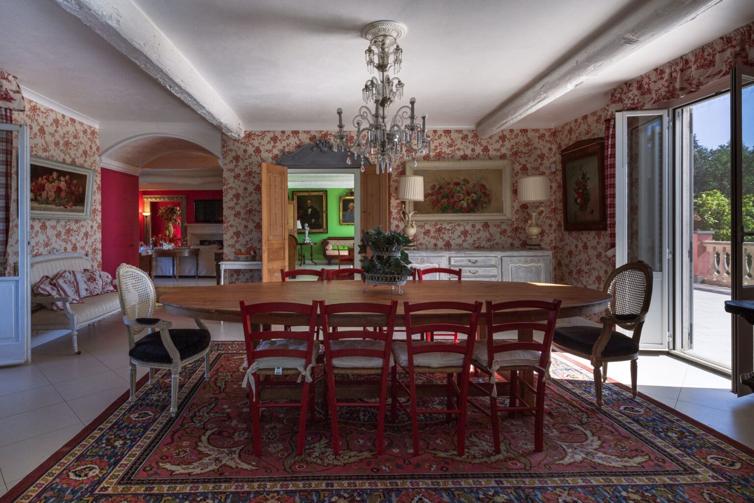 Elegant dining room featuring a large wooden table surrounded by red chairs, adorned with a decorative chandelier. The walls are decorated with floral wallpaper, complemented by classical artwork and a cozy seating area. Natural light floods the space through open doors, showcasing a vibrant outdoor view. Ideal for family gatherings and entertaining guests. | Only on SopranoVillas