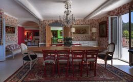 Elegant dining room featuring a large wooden table surrounded by red chairs, adorned with a decorative chandelier. The walls are decorated with floral wallpaper, complemented by classical artwork and a cozy seating area. Natural light floods the space through open doors, showcasing a vibrant outdoor view. Ideal for family gatherings and entertaining guests.