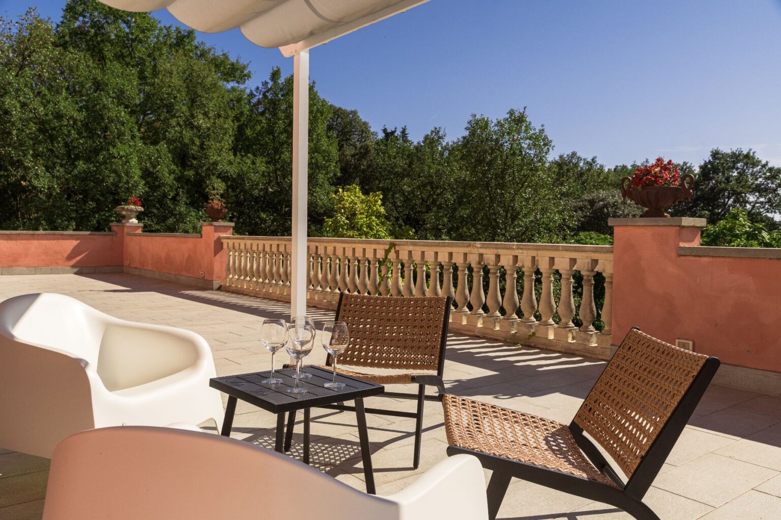 Spacious outdoor terrace featuring modern white seating, a black table with wine glasses, and a scenic view of lush greenery and decorative planters, perfect for relaxation and entertaining. | Only on SopranoVillas