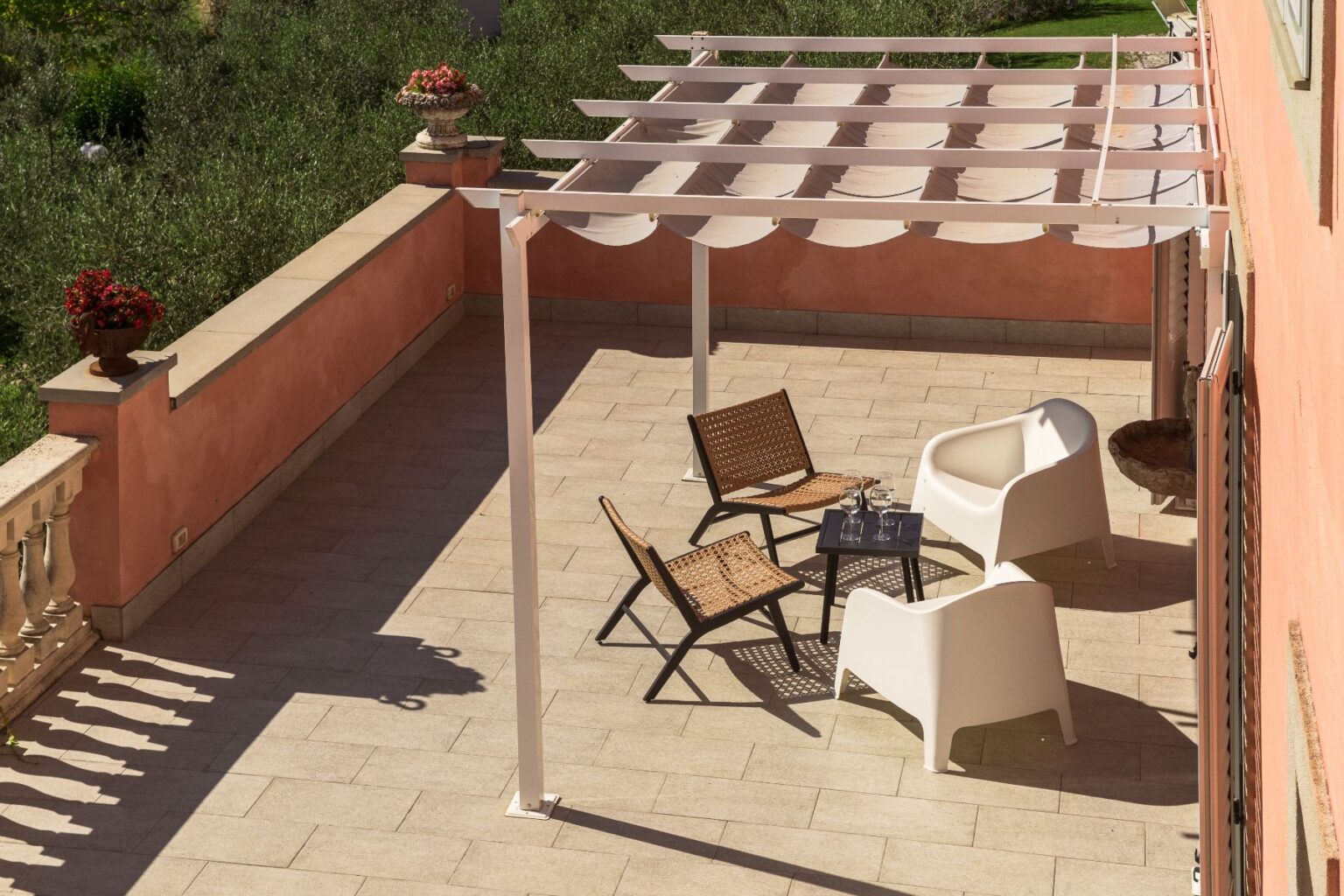 Stylish outdoor terrace featuring a modern pergola, two woven chairs, and a sleek white chair around a small table, surrounded by lush greenery and decorative flower pots. Perfect for relaxation and entertaining. | Only on SopranoVillas