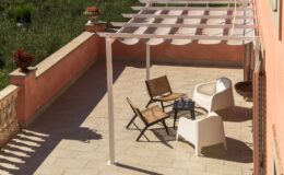 Stylish outdoor terrace featuring a modern pergola, two woven chairs, and a sleek white chair around a small table, surrounded by lush greenery and decorative flower pots. Perfect for relaxation and entertaining.