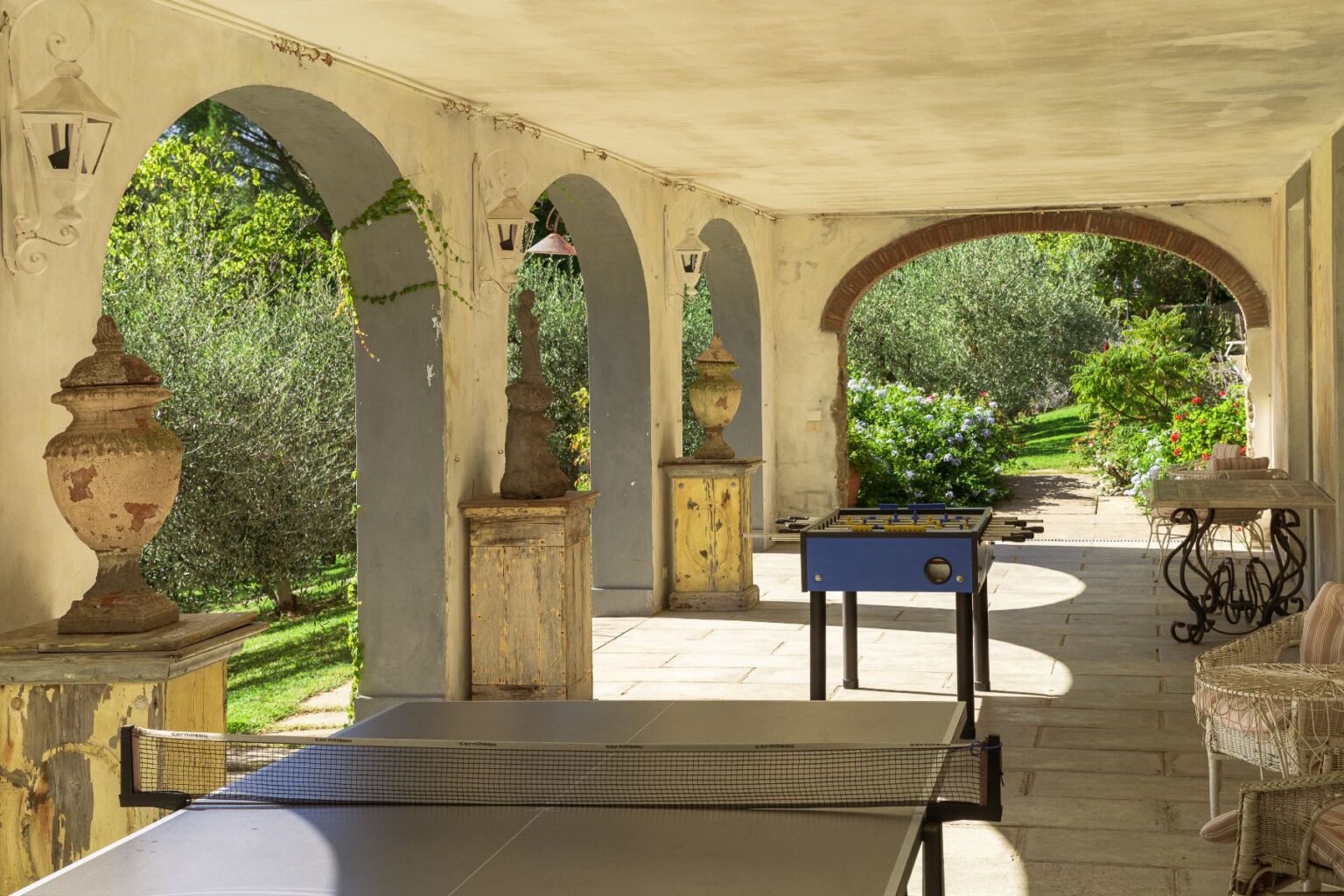 Outdoor patio with arches featuring decorative urns, a ping pong table, and a foosball table, surrounded by lush greenery and flowers in a serene garden setting. | Only on SopranoVillas