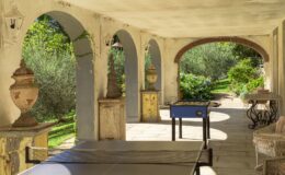 Outdoor patio with arches featuring decorative urns, a ping pong table, and a foosball table, surrounded by lush greenery and flowers in a serene garden setting.