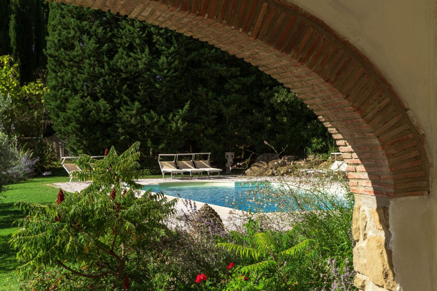Serene view of a swimming pool surrounded by lush greenery and elegant lounge chairs, framed by a rustic brick archway, creating a peaceful outdoor retreat. | Only on SopranoVillas