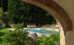 Serene view of a swimming pool surrounded by lush greenery and elegant lounge chairs, framed by a rustic brick archway, creating a peaceful outdoor retreat.
