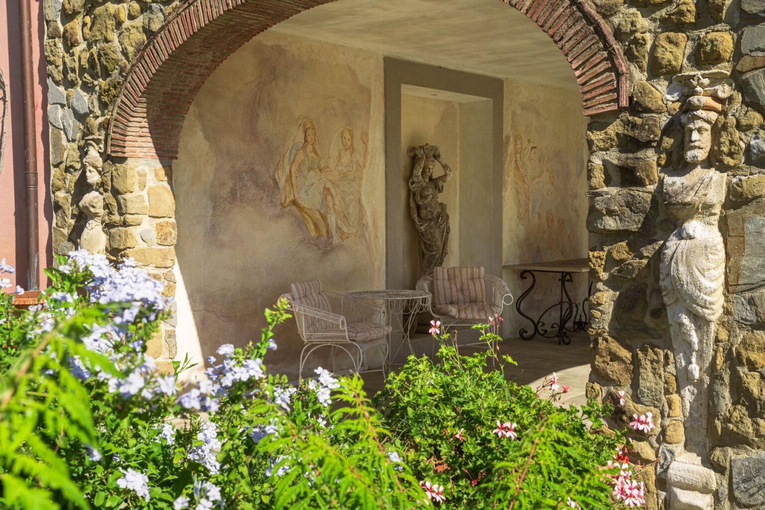 Charming outdoor patio featuring stone walls, decorative sculptures, and elegant seating surrounded by colorful flowers, showcasing artistic murals on the walls for a serene garden atmosphere. | Only on SopranoVillas