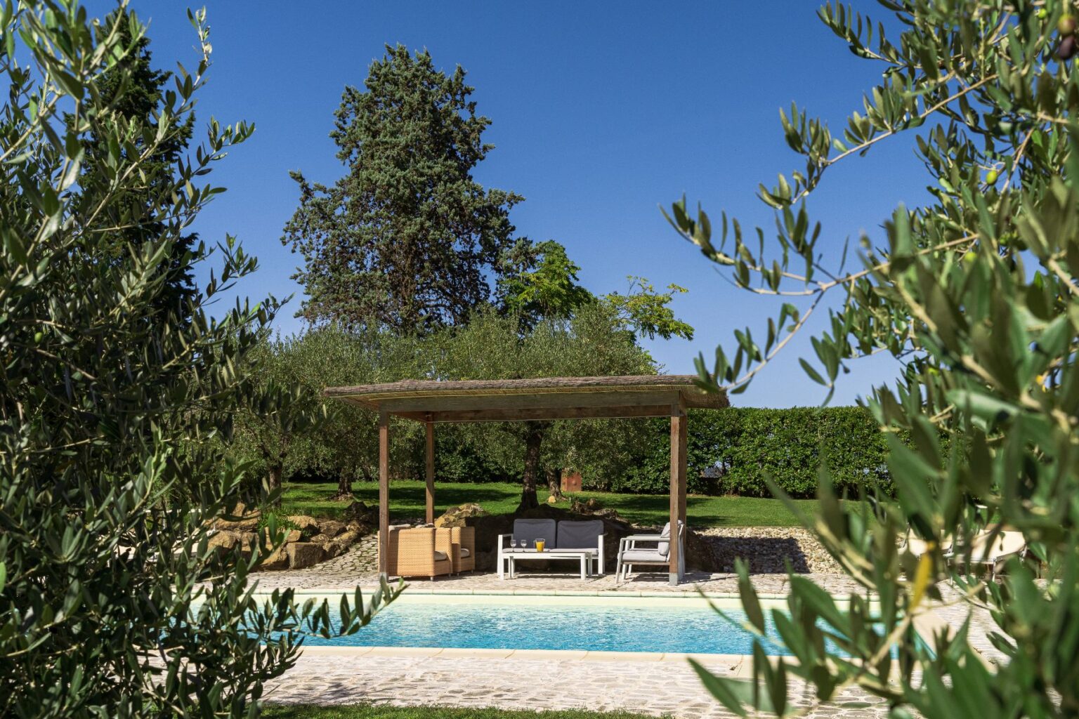 Serene outdoor pool area surrounded by olive trees, featuring a shaded seating space with comfortable furniture, perfect for relaxation in a sunny garden setting. | Only on SopranoVillas