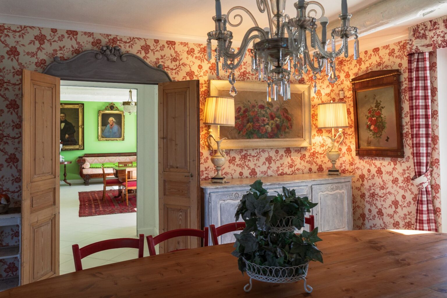 Cozy dining room with wooden table and red chairs, featuring floral wallpaper and a chandelier. An open doorway leads to a green room with vintage portraits and a patterned rug, showcasing classic decor elements. | Only on SopranoVillas