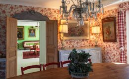 Cozy dining room with wooden table and red chairs, featuring floral wallpaper and a chandelier. An open doorway leads to a green room with vintage portraits and a patterned rug, showcasing classic decor elements.