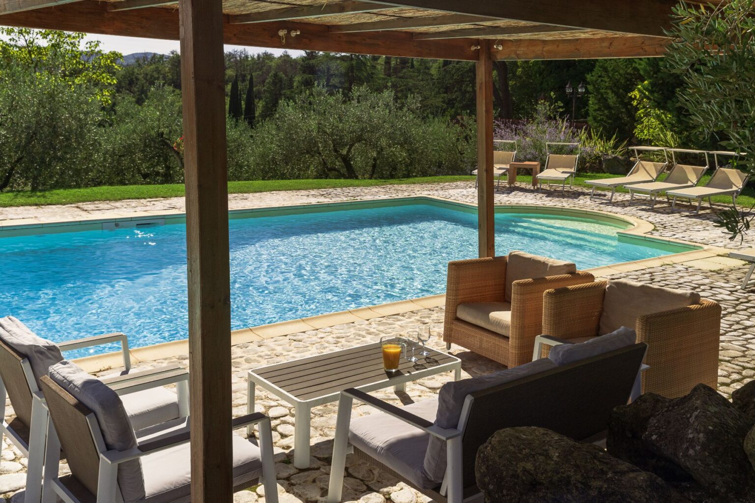 A serene outdoor pool area featuring a clear blue pool surrounded by comfortable seating and a stone patio, set against a backdrop of lush greenery and olive trees, ideal for relaxation and leisure. | Only on SopranoVillas