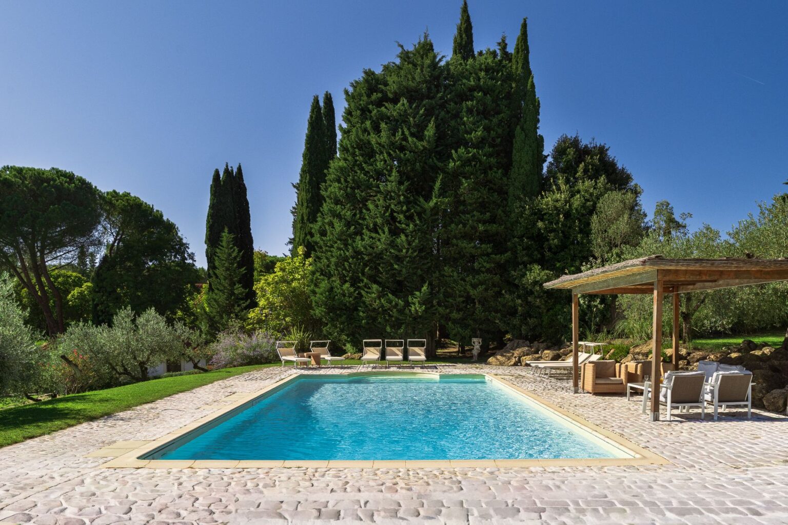 Serene outdoor pool surrounded by lush greenery and tall trees, featuring comfortable lounge chairs and a shaded seating area, perfect for relaxation in a tranquil setting. | Only on SopranoVillas