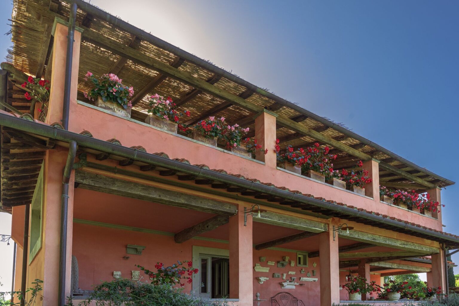 Two-story house with a pink exterior and a wooden roof, adorned with vibrant flower pots on the balcony. The structure features rustic wooden beams and a serene outdoor space, surrounded by greenery. Bright blue sky enhances the picturesque setting. | Only on SopranoVillas