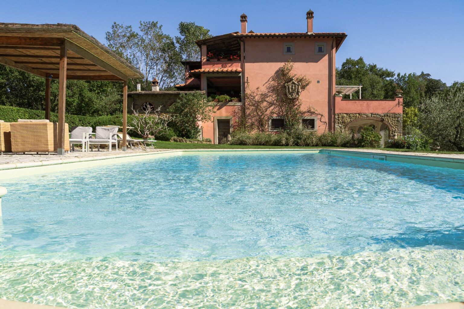 Luxurious villa with a swimming pool surrounded by lush greenery and outdoor seating, showcasing a sunny day and a charming rustic architecture. Perfect for relaxation and leisure. | Only on SopranoVillas