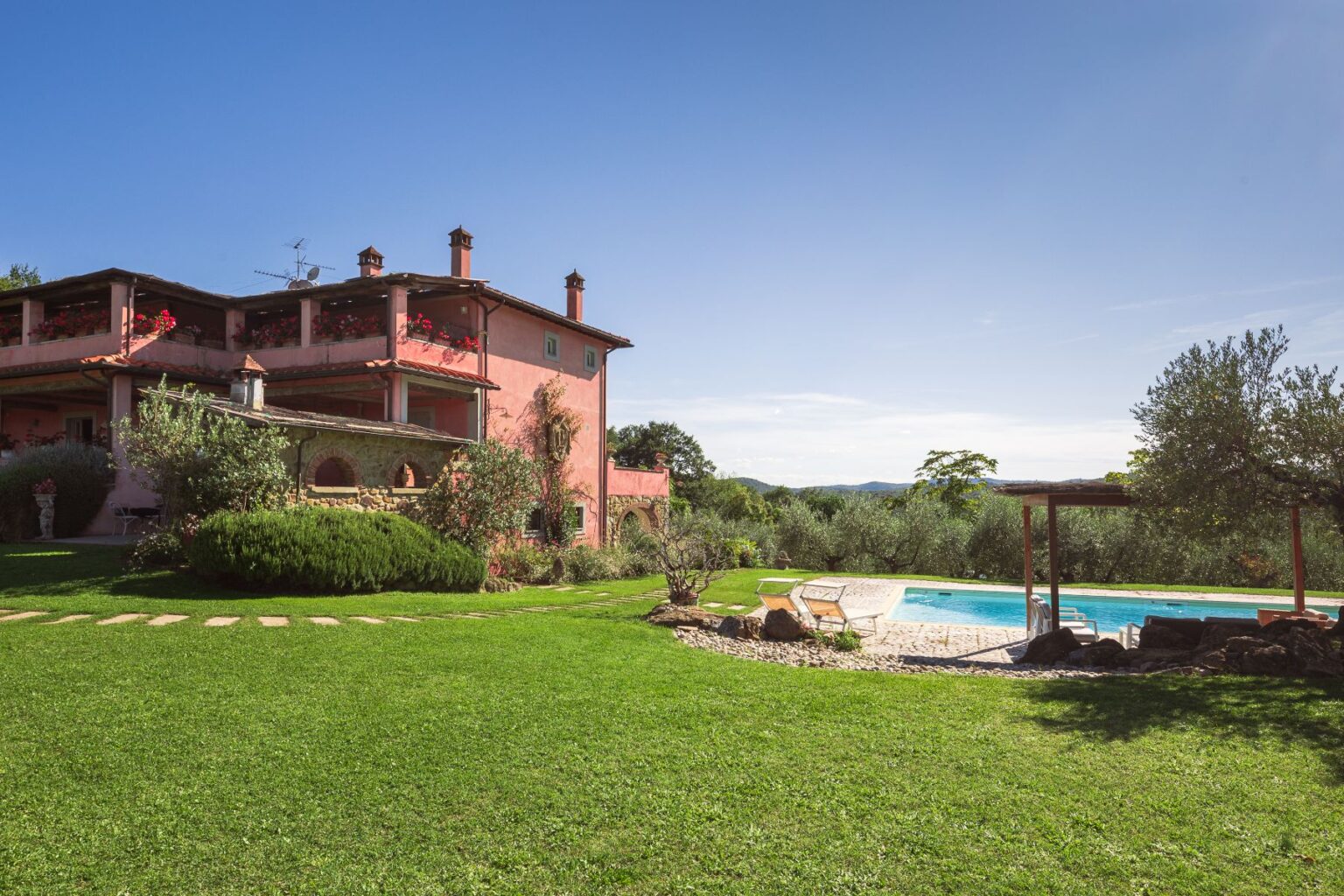 Charming pink villa with a spacious garden and swimming pool, surrounded by olive trees and lush greenery under a clear blue sky. Ideal for relaxing getaways and outdoor activities. | Only on SopranoVillas