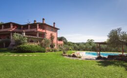 Charming pink villa with a spacious garden and swimming pool, surrounded by olive trees and lush greenery under a clear blue sky. Ideal for relaxing getaways and outdoor activities.
