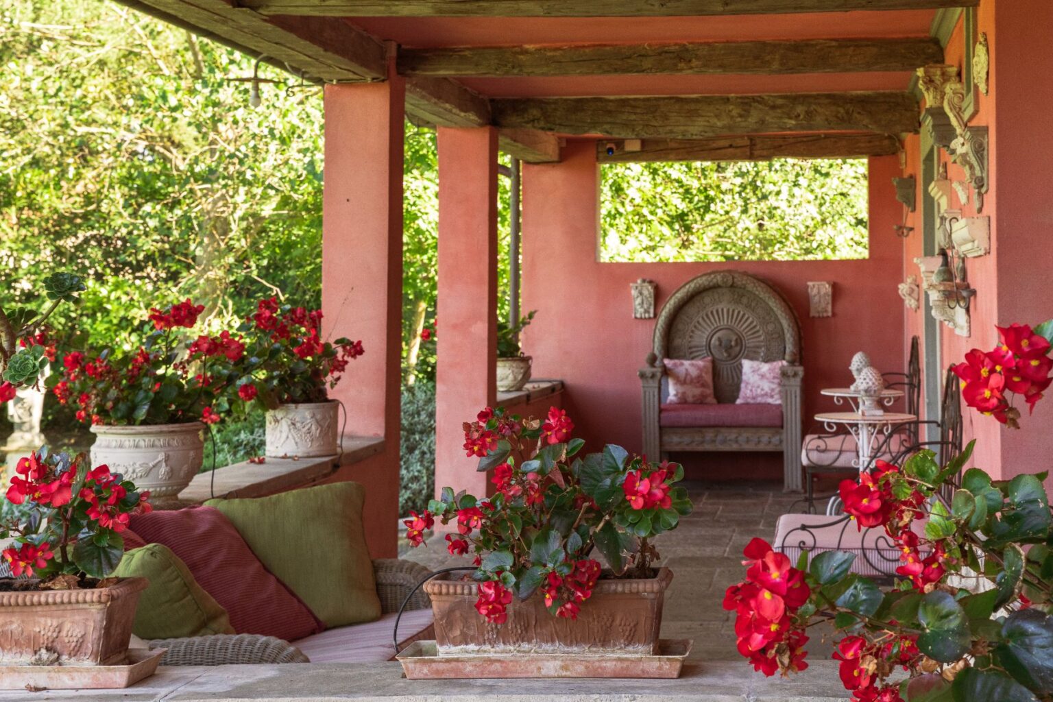 Charming outdoor patio featuring vibrant red begonias in decorative pots, plush cushions, and a beautifully carved wooden bench against a backdrop of lush greenery. Ideal for relaxation and outdoor living. | Only on SopranoVillas