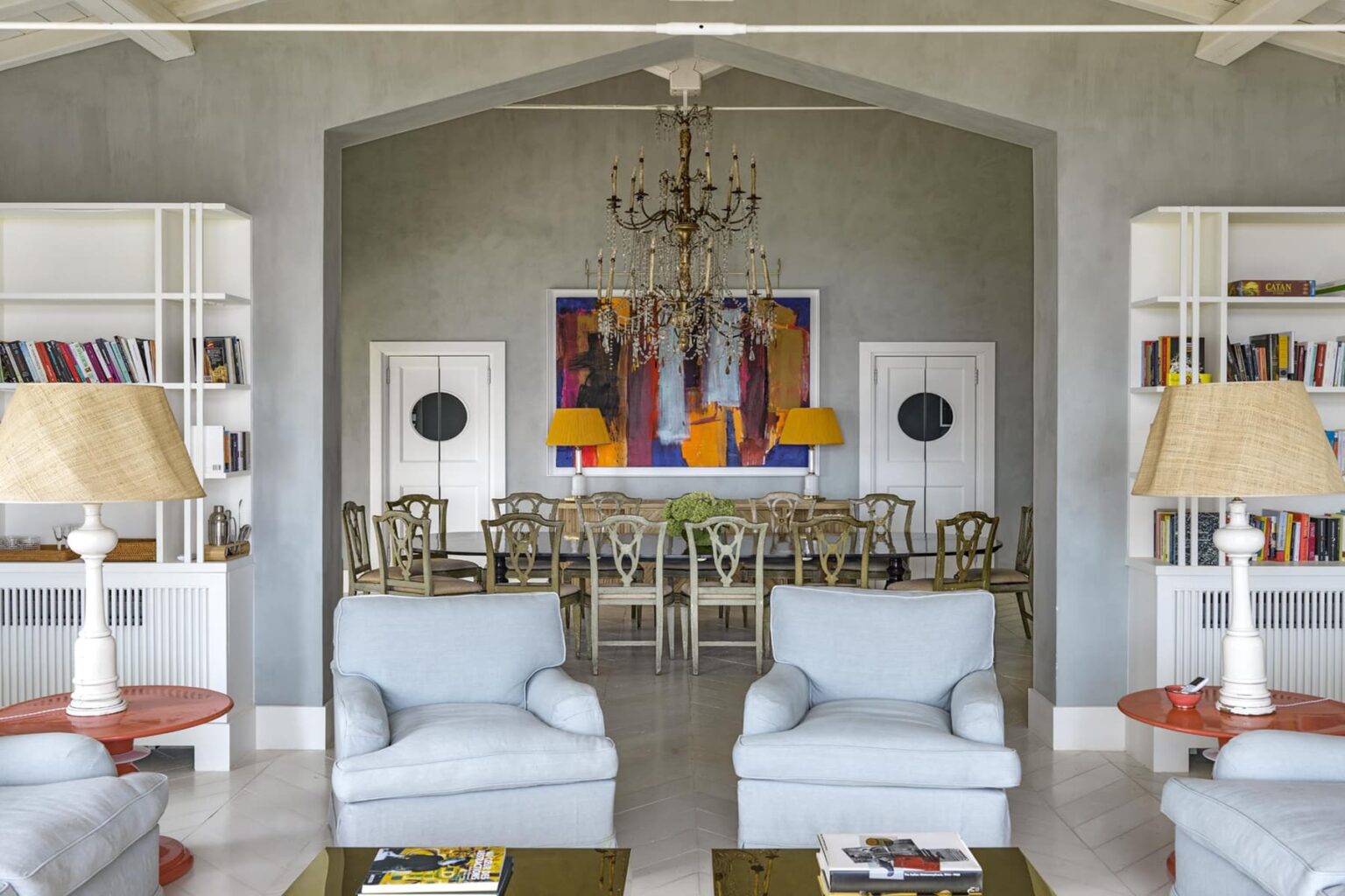 Elegant living room featuring light blue armchairs, a wooden dining table, and vibrant artwork. The space is illuminated by a chandelier and complemented by stylish lamps, with built-in bookshelves in the background. | Only on SopranoVillas